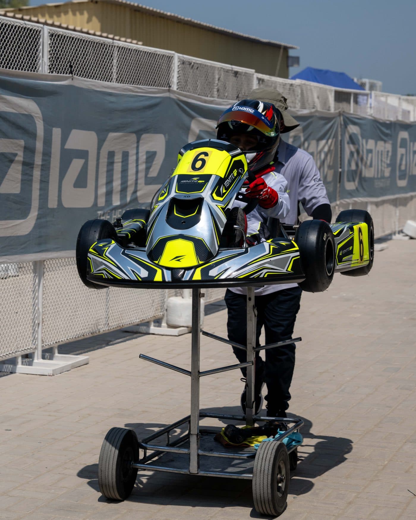 Bambino driver wheeling the kart through the paddock on its trolley, banners along the fence